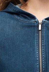 Suumärkusega sinise teksajaki poolenisti lukustatud detail, nähtav metallist lukk ja oranzhitud õmblused inimese kaela lähedal.