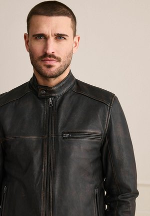 Mann mit kurzen Haaren und Bart, der eine schwarze Lederjacke mit Reißverschluss und einer Reißverschlusstasche auf der Brust vor schlichtem Hintergrund trägt.