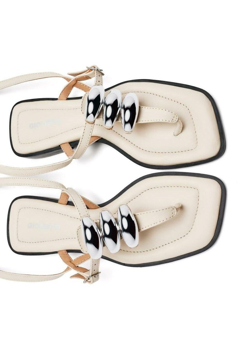 Chanclas planas beige con punta abierta, que cuentan con tres acentos plateados brillantes. Correas de cuero suave y forma cuadrada. Suela negra.