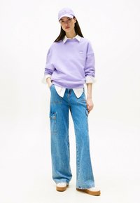 Sudadera de lavanda con cuello redondo sobre una camisa blanca de cuello, combinada con vaqueros de pierna ancha en azul claro con bolsillos de cargo y zapatos de plataforma en color beige.