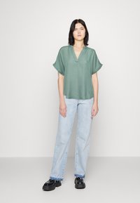 Grön kortärmad blus med V-hals och lös passform, tillsammans med ljusblå raka jeans och svarta chunky skor. Slät textur.