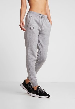 Woman wearing grey Under Armour joggers and black Under Armour sneakers, standing with hands in pockets against white background.