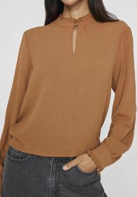 Brun blus med lång ärm och hög krage, detalj med nyckelhål och vågiga manschetter, tillverkad av textil med struktur. Matchad med mörka denim.