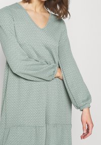 Robe vert clair avec une texture en chevron, décolleté en V et manches longues bouffantes. Dotée d'un ourlet en couches. Tissu doux.