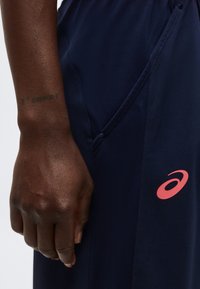 Χέρι με σκουρόχρωμο δέρμα που ακουμπάει δίπλα σε σκούρο μπλε παντελόνι με ροζ λογότυπο ASICS κοντά στη ραφή της τσέπης.