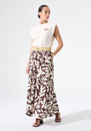 Top smanicato color crema, gonna lunga marrone con motivi di foglie bianche, vita annodata con una cintura intrecciata e sandali marroni con tacco.
