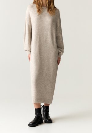 Frau trägt ein langes beiges Strickpulloverkleid mit gerippten Bündchen und Saum, kombiniert mit schwarzen Knöchelstiefeln, steht vor einem schlichten Hintergrund.