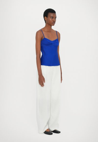 Camisola di raso blu con spalline sottili, caratterizzata da un design aderente e un dettaglio arricciato sul seno; abbinata a pantaloni bianchi larghi e ballerine nere.