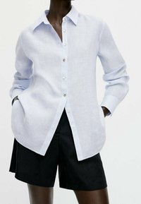 Lichtblauw gestreept overhemd met knopen, een kraag, lange mouwen en een gesplitste zoom, gecombineerd met zwarte shorts. De knopen hebben een contrasterende kleur.
