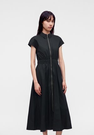 Femme portant une robe midi noire à manches courtes avec une fermeture éclair devant et une taille cintrée, debout sur un fond uni.