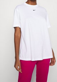 T-shirt bianca in cotone con maniche corte, scollatura rotonda e un piccolo logo nero della Nike sul petto. Abbinata a pantaloni rosa.