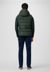 Gilet puffa verde con cappuccio, design aderente, indossato sopra una maglietta a maniche lunghe scura, abbinato a pantaloni blu navy e scarpe scure.