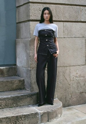 Joven mujer que lleva una camiseta gris debajo de un corsé vaquero negro con botones y jeans anchos a juego, de pie en unos escalones de piedra al aire libre.