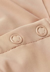 Detailaufnahme eines beigen Stoffes mit zwei runden Knöpfen, glatter Textur und sichtbaren Nähten entlang der Ränder.