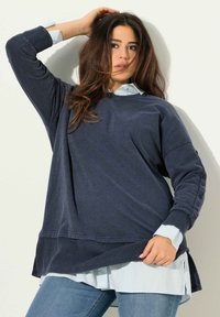 Felpa oversize blu scuro con polsini a costine, caratterizzata da bottoni decorativi sulle maniche, indossata sopra una camicia col collo azzurra.