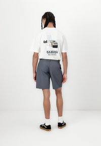 Hvit grafisk t-skjorte med trykk på ryggen som viser en lastebil og tekst, kombinert med grå shorts. Modellen har på seg svarte joggesko og hvite sokker.