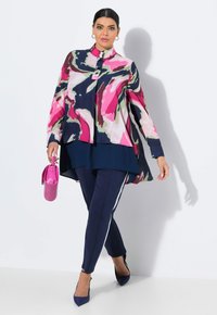 Blouse à imprimé floral avec un design asymétrique en rose, marine et vert ; associée à un pantalon marine avec des rayures blanches sur les côtés et un sac à main rose.