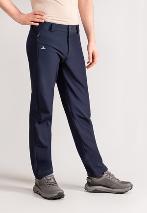 Granatowe spodnie softshell o slim fit, z zamkniętą kieszenią na zamek, paskami na pasek i subtelnym logo. W zestawie z szarymi butami trekkingowymi.