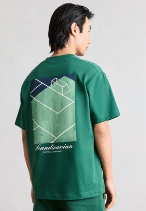Férfi zöld pólóban, amelyen egy grafikus futballpálya és a "Scandinavian Football Community" felirat van a hátán, oldalra néz.