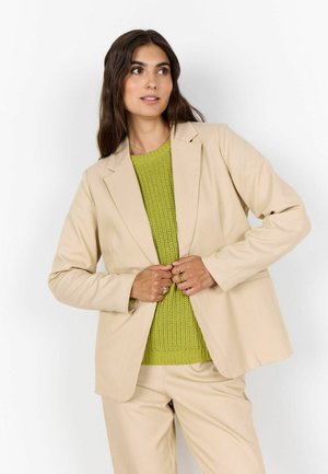 Frau mit langen dunklen Haaren, die einen beigen Anzug und einen grünen Strickpullover trägt, richtet die Knöpfe ihrer Jacke und blickt leicht zur Seite.