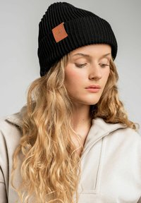 Czarna, prążkowana czapka beanie z skórzaną naszywką, charakteryzująca się ciasnym dopasowaniem i teksturowaną powierzchnią. Noszona z beżową bluzką i ułożona z falującymi włosami.