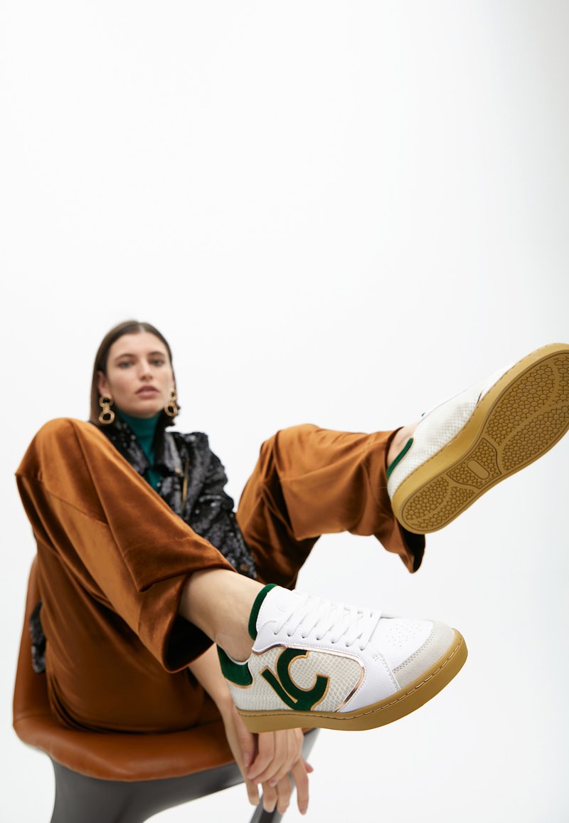 Weiße Sneaker mit grünen Akzenten und текстuriertem Material. Verfügt über eine Gummisohle und ein gemustertes Seitenlogo. Das Modell trägt braune Samdhosen.