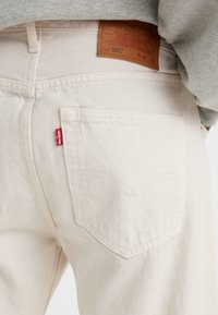 Jeans de denim color crema con un diseño clásico de cinco bolsillos, que cuentan con una etiqueta de cuero marrón en la cintura y una etiqueta de logo roja en el bolsillo trasero.