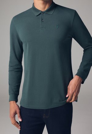 Homme portant un polo manches longues vert foncé avec deux boutons et un petit logo brodé sur la poitrine gauche, associé à un jean foncé.