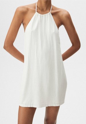 Ærmeløs hvid halterneck-kjole med løs pasform, båret af en person, der står med hænderne bag ryggen.