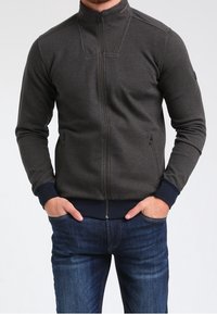Giacca a zip grigio scuro con motivo testurizzato, colletto alto e polsini a costine blu. Due tasche laterali con chiusura a zip. Indossata sopra jeans blu.