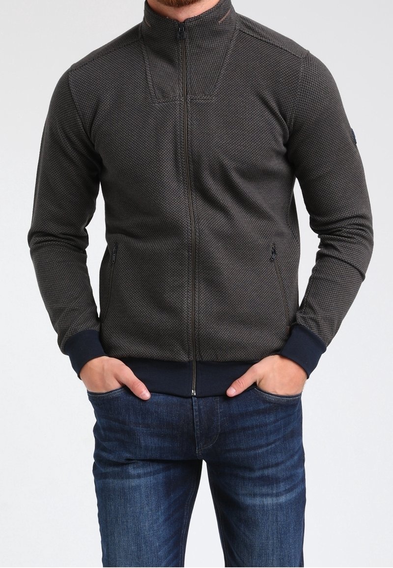 Giacca a zip grigio scuro con motivo testurizzato, colletto alto e polsini a costine blu. Due tasche laterali con chiusura a zip. Indossata sopra jeans blu.