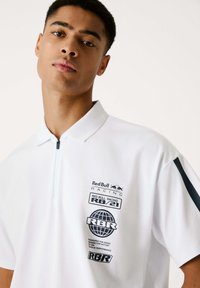 Camiseta polo blanca con cuello con cremallera, acentos en azul marino en las mangas y una marca impresa con texto y logotipos en el lado izquierdo.