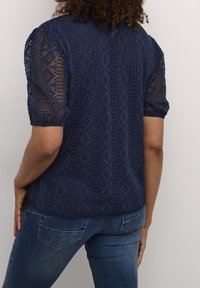 Mörkblå topp med diamantmönstrad spetsdetalj och korta puffärmar, parat med denimjeans. Texturerad tyg med en halvgenomskinlig effekt.