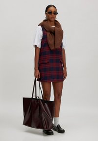 Gestreiftes Kleid in Marineblau und Rot mit einem braunen Strickschal, schwarzen Sonnenbrillen und einer großen glänzenden burgunderfarbenen Tote-Bag. Weißes T-Shirt und Socken, schwarze Schuhe.