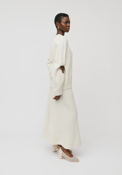 Kreminė kabelinė megzta svarkas, ilga kreminė sijonas ir šviesiai smėlio spalvos blokiniai kulniukai. Sklandus paviršius su laisvu siluetu ir elegantiška forma.