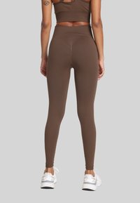 Kobieta w brązowych legginsach z wysokim stanem i dopasowanym biustonoszu sportowym, stoi z nogami na szerokość barków w białych sneakersach, zwrócona tyłem.