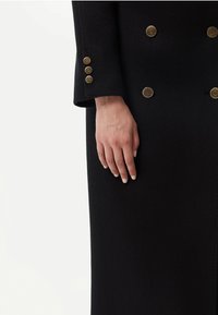 Svart ullkappa med guldfärgade dekorativa knappar på ärmarna och framsidan, med en skräddarsydd passform och slät textur.