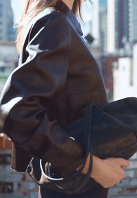 Czarna skórzana krótka kurtka o gładkiej fakturze z podwiniętymi rękawami. Torebka trzymana w ręku wykonana jest z ciemnego zamszu o miękkiej powierzchni i minimalistycznych metalowych elementach.