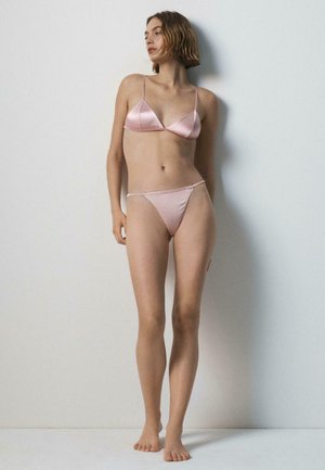 Jeune femme aux cheveux courts, debout pieds nus contre un mur clair, portant un soutien-gorge en satin rose et une culotte assortie, regardant sur le côté.