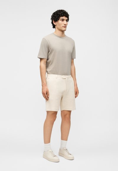 Άνδρας στέκεται φορώντας ανοιχτό μπεζ σορτς, γκρι κοντομάνικο μπλουζάκι, άσπρες κάλτσες και μπεζ αθλητικά παπούτσια σε απλό φόντο.