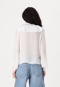 Weiße, texturierte Bluse mit einem Rüschensaum und langen puffärmel, kombiniert mit hellblauen Denim-Shorts mit Gesäßtaschen.