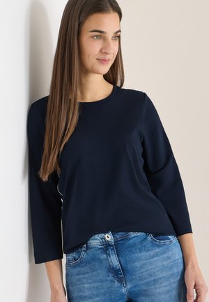 Femme aux longs cheveux bruns portant un haut à manches longues bleu marine et un jean bleu, appuyée contre un mur blanc, regardant sur le côté.