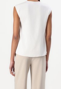 Blouse blanche sans manches avec un col rond, tissu lisse, coupe décontractée, ourlet droit et accents aux épaules, associée à un pantalon beige.