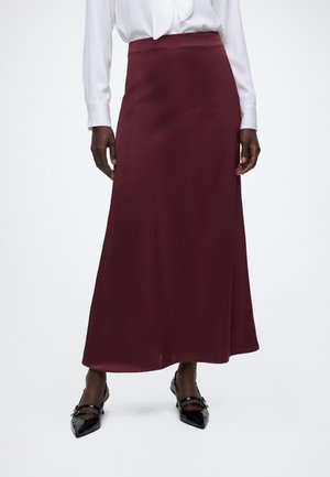 Femme portant un chemisier blanc à manches longues et une jupe bordeaux taille haute jusqu'aux chevilles avec des chaussures noires pointues.