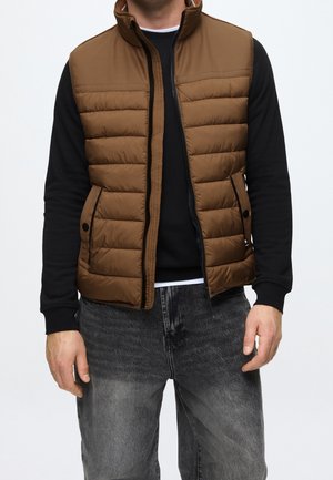 Mand iført en brun polstret vest over en sort langærmet skjorte og falmede sorte jeans, stående mod en ensfarvet baggrund.