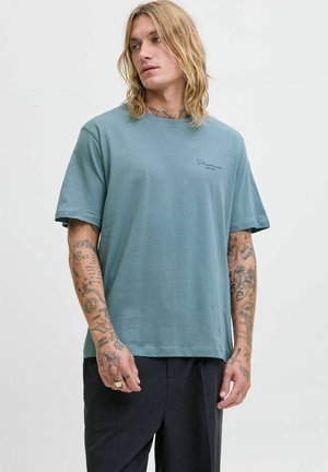 Blond man med långt hår som bär en blå "Premium" t-shirt och mörka byxor, visar tatuerade armar och tittar åt sidan.