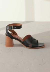 Schwarze Leder-Absatzsandalen mit Überkreuzriemen-Design, verstellbarem Knöchelriemen, braunem Holzblockabsatz und offener Zehenpartie.