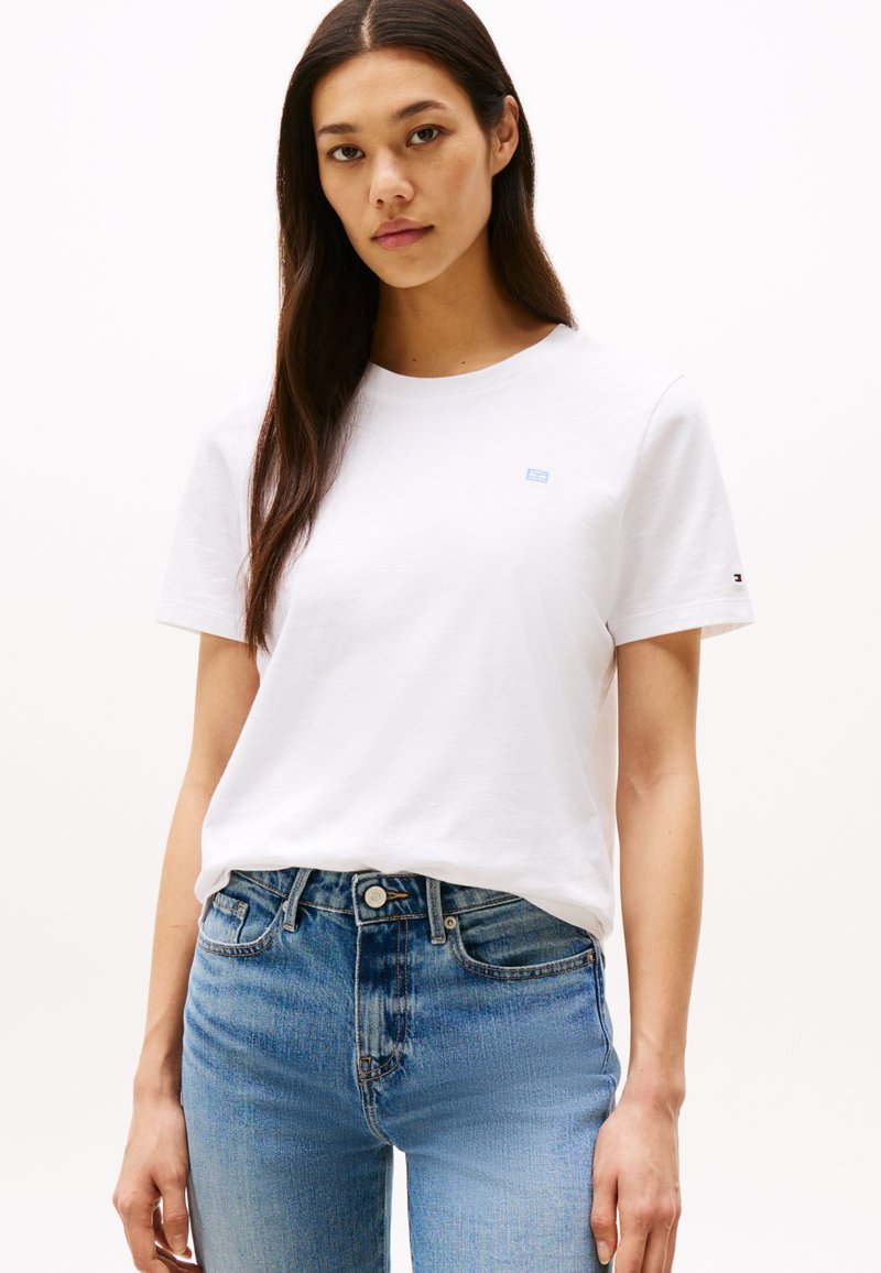 Camiseta blanca de algodón con cuello redondo, mangas cortas y un logo azul claro en el pecho; combinada con jeans de mezclilla azul claro.