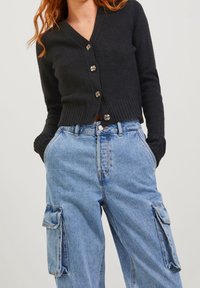 Cardigan nero a coste con scollatura a V e chiusura a cinque bottoni, abbinato a jeans cargo blu chiaro con tasche laterali.