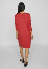 Robe tricotée rouge à manches longues, avec des poignets côtelés et un ourlet arrondi. Texture lisse, design mi-long, accompagnée de sandales plates noires.